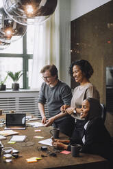 Smiling male and female colleague standing by businesswoman sitting at conference table in meeting room - MASF30987