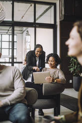 Smiling businesswoman discussing over laptop with female colleague sitting on chair in creative office - MASF30996