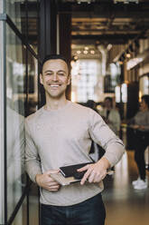 Porträt eines glücklichen reifen Geschäftsmannes mit Laptop, der in einem kreativen Büro steht - MASF31009