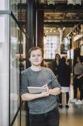 Porträt eines jungen Geschäftsmannes mit Laptop an der Wand lehnend in einem kreativen Büro - MASF31011