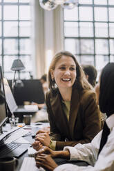 Happy mid adult businesswoman discussing with female colleague sitting at desk in office - MASF31022