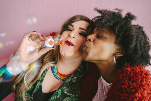Non-binary person with female friend blowing bubbles against wall - MASF31047