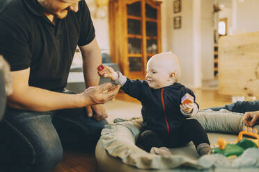 Smiling male toddler giving toy block to father sitting in living room at home - MASF31067