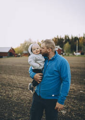 Father kissing toddler son standing at farm during sunset - MASF31087