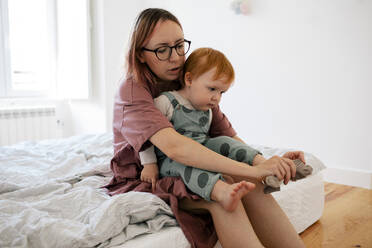 Mother puts sock to male toddler's foot sitting in bedroom - MASF31095