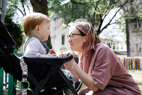 Mother making faces looking at son sitting in baby stroller - MASF31101