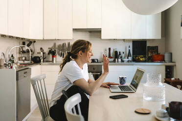 Happy female freelancer waving on video call through laptop at table in kitchen - MASF31108