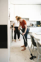 Woman cleaning floor with vacuum cleaner while girlfriend standing in kitchen - MASF31117