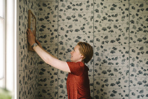 Side view of woman adjusting picture frame on wall at home - MASF31127