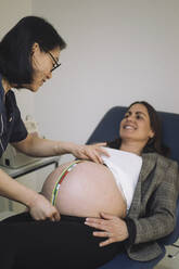Smiling doctor measuring abdomen of pregnant patient lying on examination table in medical clinic - MASF31159