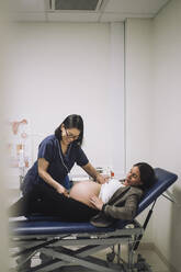 Smiling gynecologist measuring abdomen of pregnant woman lying on gurney in hospital - MASF31160