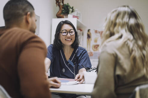 Happy female healthcare expert discussing over chart with couple in medical clinic - MASF31199