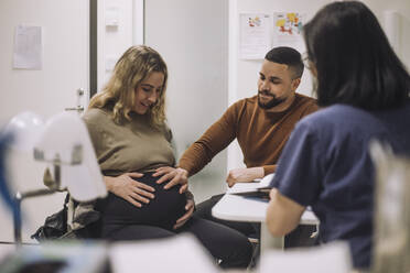 Lächelnder Mann, der den Bauch einer schwangeren Frau berührt, während er mit einem Gynäkologen in einer medizinischen Klinik spricht - MASF31208