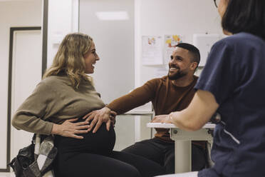 Glücklicher Mann, der den Bauch einer schwangeren Frau berührt, während er mit einer Ärztin in einer medizinischen Klinik sitzt - MASF31209