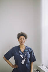 Portrait of happy female doctor standing with hand on hip against white wall in hospital - MASF31228