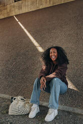 Happy young woman with bag sitting on slope at roadside - MASF31249