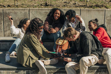 Happy man applying nail polish on hand of woman while sitting by cheerful friends in park - MASF31266