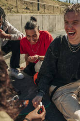 Woman painting fingernail of happy man sitting by friends in park on sunny day - MASF31267