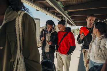 Junger Mann teilt sein Smartphone mit einem männlichen Freund, während er auf dem Bahnsteig spazieren geht - MASF31299