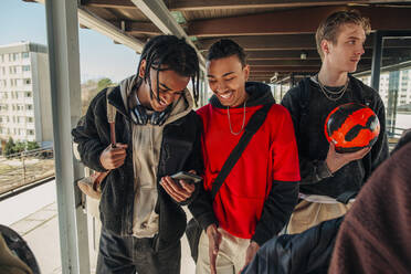 Glückliche männliche Freunde teilen sich ein Smartphone beim Spaziergang am Bahnhof - MASF31300