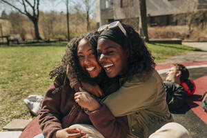Glückliche multirassische Freundinnen, die sich gegenseitig umarmen, während sie auf einem Spielplatz an einem sonnigen Tag sitzen - MASF31346