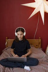 In voller Länge der konzentrierten Teenager-Mädchen mit dunklen Mädchen in Freizeitkleidung und Kopfhörer sitzen auf bequemen Bett mit gekreuzten Beinen und Tippen auf dem Laptop, während die Hausaufgaben Zuordnung - ADSF35474
