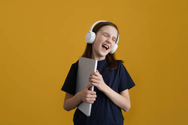 Positives Teenager-Mädchen mit dunklen Haaren im T-Shirt, das mit geschlossenen Augen ausdrucksvoll singt, während es mit Kopfhörern Musik hört und mit einem Laptop in den Händen vor einem gelben Hintergrund steht - ADSF35478