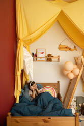 Adorable kleines Kind mit langen dunklen Haaren liegen auf dem Bett unter warmen Decke und Musik hören in Kopfhörer in gemütlichen Schlafzimmer mit Baldachin dekoriert - ADSF35488