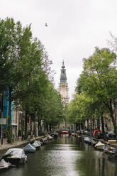 Malerische Kulisse der alten Zuiderkerk-Kirche in der Nähe einer ruhigen Gracht mit vertäuten Booten inmitten von Wohngebäuden an einem bewölkten Tag in Amsterdam - ADSF35493