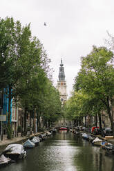 Picturesque scenery of aged Zuiderkerk church located near calm canal with moored boats amidst residential buildings on cloudy day in Amsterdam - ADSF35493