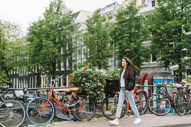 Volle Länge der positiven jungen hispanischen weiblichen Reisenden mit langen dunklen Haaren in stilvollem Outfit zu Fuß auf gepflasterten Fußgängerbrücke in der Nähe geparkten Fahrrädern während Sightseeing-Tour in Amsterdam - ADSF35494
