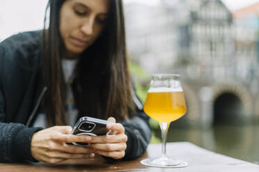 Erfreute junge ethnische Dame mit langen dunklen Haaren in Jacke lächelt glücklich, während sie an einem Holztisch mit einem Glas Bier sitzt und auf einem Handy in der Nähe einer Gracht in Amsterdam Nachrichten schreibt - ADSF35495