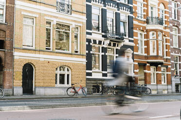 Unbekannte Person auf dem Fahrrad auf der Straße in der Nähe von alten Backstein-Wohngebäuden in sonnigen Morgen in Amsterdam - ADSF35498