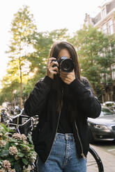 Unerkennbare junge Touristin mit langen dunklen Haaren in modischer Jacke, die ein Foto mit einem Fotoapparat macht, während sie auf der Straße in der Nähe eines geparkten Fahrrads steht, während eines Urlaubs in Amsterdam - ADSF35500