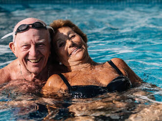 Cheerful senior couple smiling brightly while swimming together in outdoor pool during summer holidays - ADSF35501