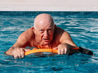 Aktiver älterer männlicher Tourist übt Schwimmen mit Schwimmbrett im Freibad und schaut in die Kamera an einem sonnigen Tag - ADSF35503