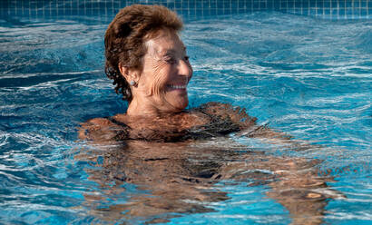 Fröhliche ältere Frau in Badekleidung lächelt fröhlich und schaut weg, während sie an einem sonnigen Tag im Pool schwimmt - ADSF35504