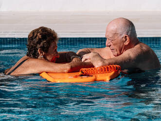 Seitenansicht einer glücklichen älteren Frau und eines glücklichen älteren Mannes, die sich beim Schwimmen im Freibad an einem sonnigen Tag auf ein Kickboard stützen und sich gegenseitig ansehen - ADSF35505