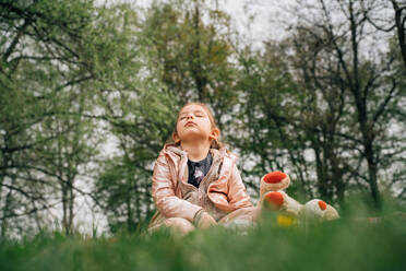 Ebenerdige Darstellung eines niedlichen kleinen Kindes in warmer Kleidung mit einem Plüschbären, der mit geschlossenen Augen auf einer Wiese vor hohen grünen Bäumen im Park ruht - ADSF35506