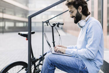 Smiling freelancer using laptop sitting by railing - XLGF02958