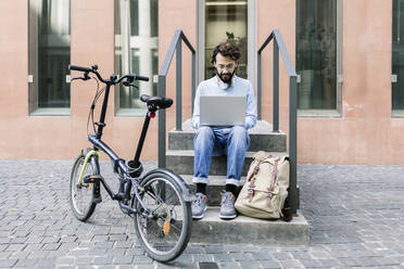 Businessman using laptop sitting on steps by bicycle - XLGF02959