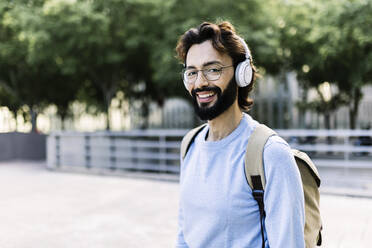 Happy bearded man with backpack wearing wireless headphones - XLGF02984