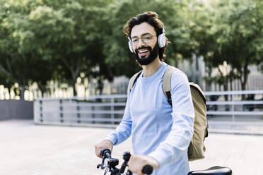 Smiling man wearing wireless headphones standing with bicycle - XLGF02985