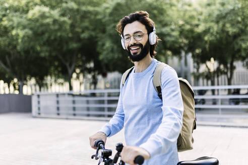 Lächelnder Mann mit kabellosen Kopfhörern, stehend mit Fahrrad - XLGF02985