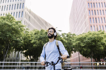 Happy man wearing wireless headphones standing with bicycle - XLGF02986