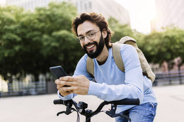Happy man using smart phone leaning on bicycle - XLGF02992