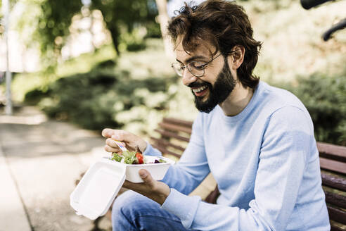 Lächelnder Mann mit Salat auf einer Bank im Park sitzend - XLGF03002