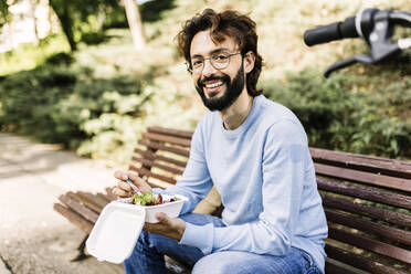 Glücklicher Mann mit Essen auf einer Bank im Park sitzend - XLGF03003