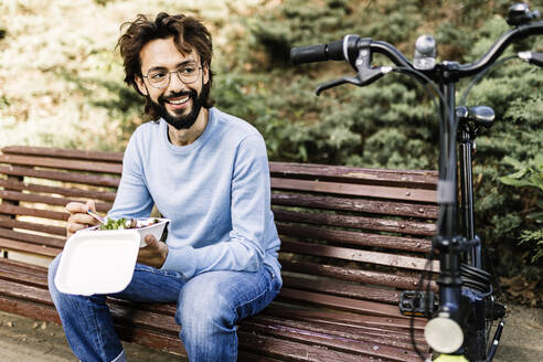 Mann mit Essen auf Parkbank sitzend mit Blick auf Fahrrad - XLGF03004