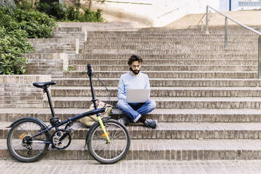 Freelancer using laptop sitting on steps near bicycle - XLGF03005